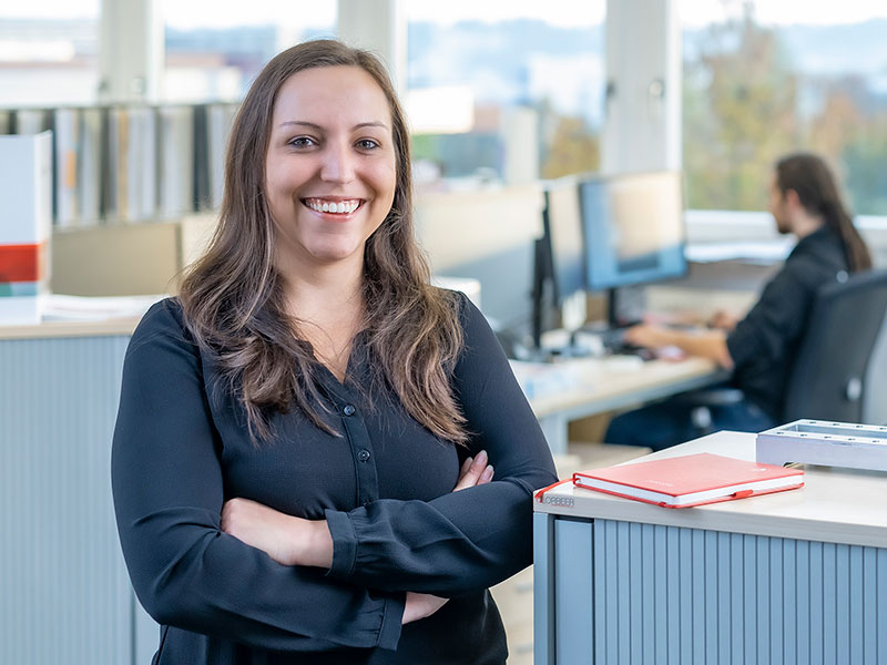 Female mployee stands in open plan office and smiles